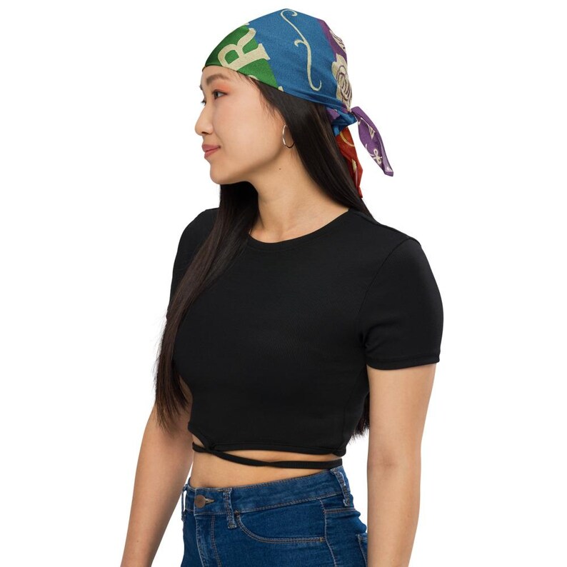 May include: A colorful bandana with a blue background and various designs, including the letter "R". The bandana is tied at the back. The person is wearing a black crop top and blue jeans.