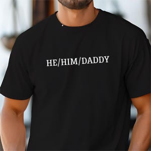 May include: Black t-shirt with white text that reads "HE/HIM/DADDY".