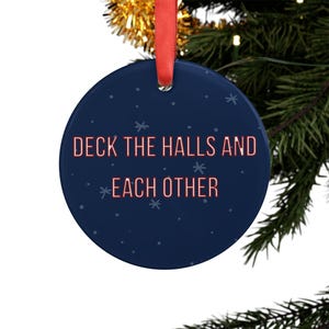 May include: A round, dark blue ceramic ornament featuring a red ribbon for hanging. The text "DECK THE HALLS AND EACH OTHER" is printed in a light red, sans-serif font across the center, surrounded by small, light blue snowflake illustrations.