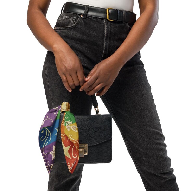 May include: A black handbag with a gold clasp and a colorful scarf tied to the handle. The scarf features a rainbow design with stars and swirls. The person is wearing black jeans and a white top.