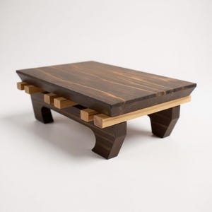 Peut inclure: Petite table rectangulaire en bois, avec un plateau teinté brun foncé et des accents brun clair. La table présente un design unique avec une structure de support à lattes et des pieds inclinés. Mesures: environ 46 cm de long, 25 cm de large et 15 cm de haut.