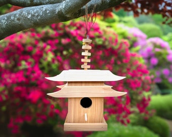 May include: A wooden birdhouse shaped like a pagoda with a white roof and a small opening for birds to enter. The birdhouse is hanging from a tree branch in a garden setting.
