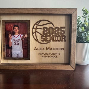 Könnte beinhalten: Ein Holzrahmen mit einem Foto eines Basketballspielers in einem weißen Trikot mit der Nummer 25. Der Rahmen enthält auch eine Basketballgrafik und den Text "2025 Senior" mit dem Namen "Alex Madden" und "Hancock County High School" darunter.
