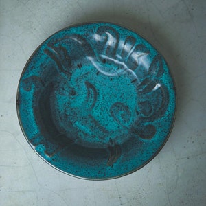 Könnte beinhalten: Eine tealblaue Keramikschüssel mit einem dekorativen Design. Die Schüssel hat eine gesprenkelte Glasur und einen dunkelblauen Rand.