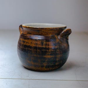 May include: A small brown ceramic pot with two handles. The pot has a dark brown glaze with a light brown stripe pattern.