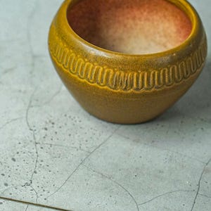 Może przedstawiać: Mała, okrągła, żółta doniczka ceramiczna z fakturowanym brzegiem. Doniczka jest pusta i ma jasnobrązowe wnętrze.
