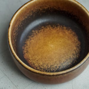 May include: A small, round ceramic bowl with a gradient of colors. The exterior is a warm brown, transitioning to a darker brown interior with a speckled, textured appearance. The bowl is empty and sits on a light-colored surface.