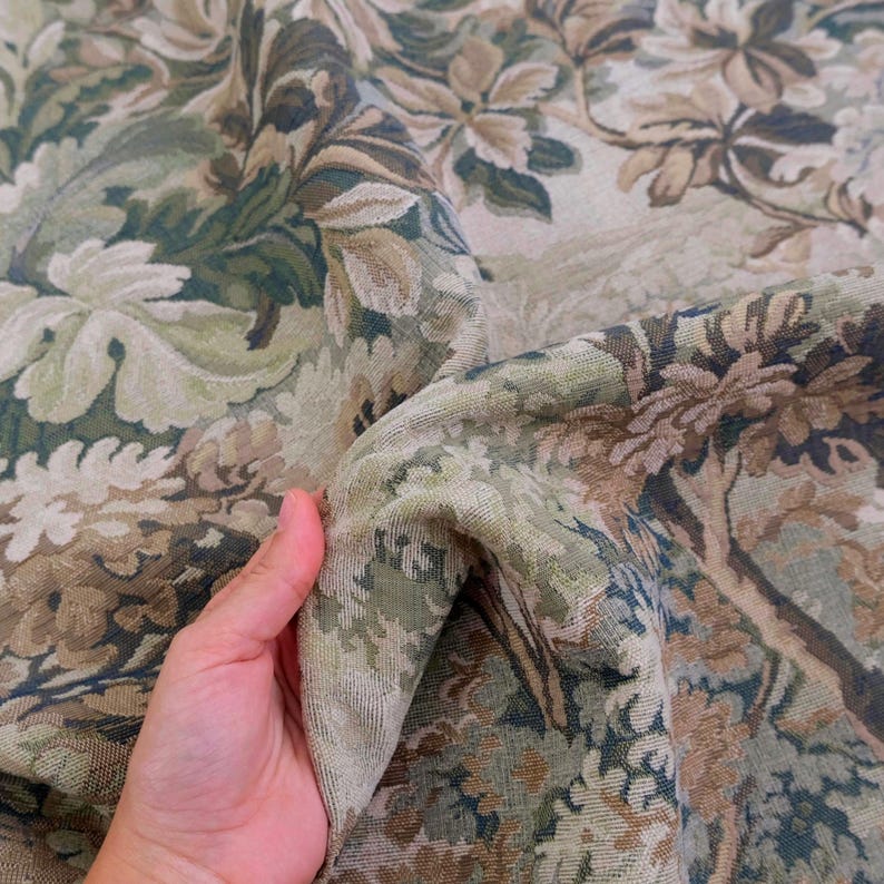 May include: A woven fabric featuring a dense, intricate botanical pattern in shades of sage green, olive, beige, and cream. The textile displays a repeating design of large, stylized leaves and floral elements with a textured, tapestry-like finish.