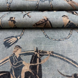 Puede incluir: Primer plano de una tela azul claro con un dise&ntilde;o ecuestre repetido. El patr&oacute;n presenta jinetes, caballos, borlas y detalles de cuerda en tonos marr&oacute;n y azul marino. La tela est&aacute; doblada, mostrando m&uacute;ltiples capas.