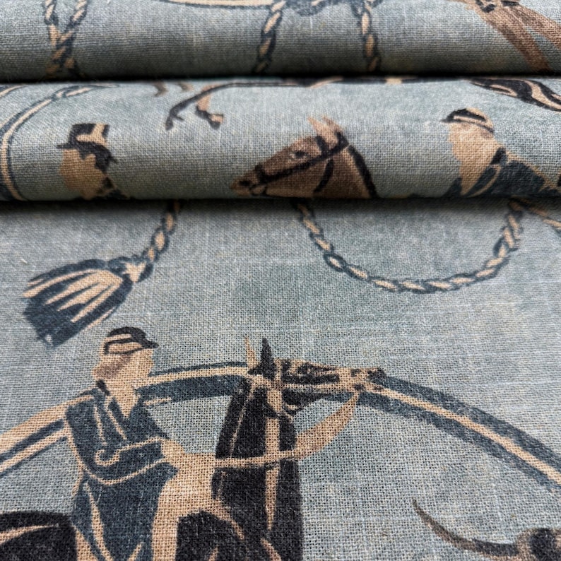 Puede incluir: Primer plano de una tela azul claro con un dise&ntilde;o ecuestre repetido. El patr&oacute;n presenta jinetes, caballos, borlas y detalles de cuerda en tonos marr&oacute;n y azul marino. La tela est&aacute; doblada, mostrando m&uacute;ltiples capas.