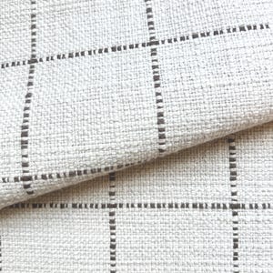 Könnte beinhalten: Nahaufnahme eines strukturierten, cremefarbenen Stoffes mit einem Gittermuster aus dunkelbraunen Linien. Der Stoff ist gefaltet und zeigt das Gewebe und die Textur. Die Gitterlinien sind gleichmäßig angeordnet und erzeugen ein modernes, geometrisches Design.