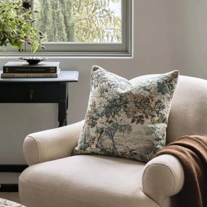 May include: A square throw pillow featuring a detailed woven tapestry design of trees and foliage in muted shades of green, blue, and brown. The pillow is placed on a cream-colored upholstered armchair, which also holds a brown throw blanket.
