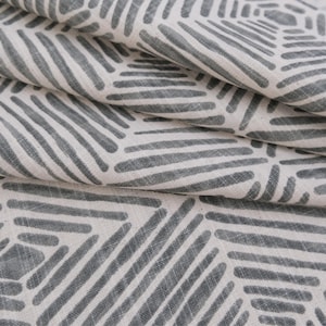 May include: Close-up of a folded fabric with a repeating pattern. The pattern consists of gray, elongated shapes arranged in diagonal rows against a white background. The fabric has a textured appearance, with visible weave details.