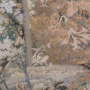 May include: A woven fabric featuring a dense, intricate pattern of foliage, trees, and floral elements in shades of sage green, muted gold, brown, and cream. The textile displays a visible vertical seam with a textured, tightly stitched edge.