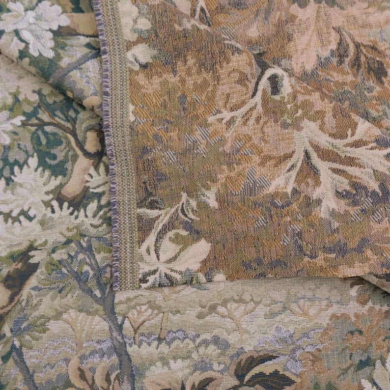 May include: A woven fabric featuring a dense, intricate pattern of foliage, trees, and floral elements in shades of sage green, muted gold, brown, and cream. The textile displays a visible vertical seam with a textured, tightly stitched edge.
