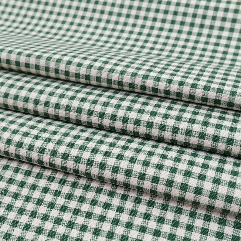 May include: A folded textile fabric featuring a classic gingham pattern with a repeating grid of dark forest green and off-white squares. The material displays a visible woven texture with consistent, uniform checks across the entire surface.