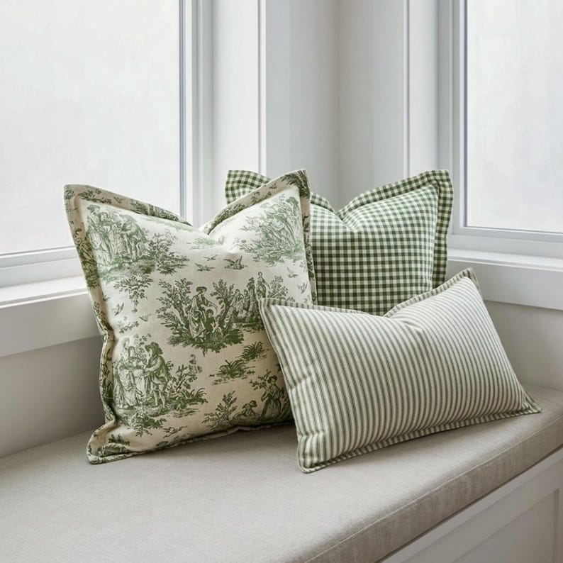 May include: Three decorative throw pillows in a window seat. One pillow features a green and white toile pattern, another has a green gingham check, and the third has green and white stripes. The pillows are arranged on a neutral-colored cushion.