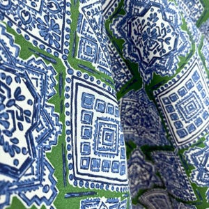 May include: Close-up of fabric with a repeating geometric pattern in blue and white on a green background. The design features squares, diamonds, and floral elements, creating a textured, layered effect. The fabric is folded, showing depth and dimension.