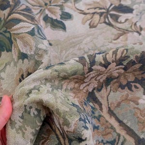 May include: A woven fabric featuring a detailed botanical pattern in shades of sage green, muted brown, and cream. The textile displays a dense, textured weave with intricate floral and leafy motifs that create a layered, multi-dimensional appearance.