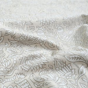 May include: A beige fabric featuring a repeating pattern of white line-drawn birds and intricate floral motifs. The textile has a visible woven texture with soft folds that create depth and shadows across the surface of the patterned material.