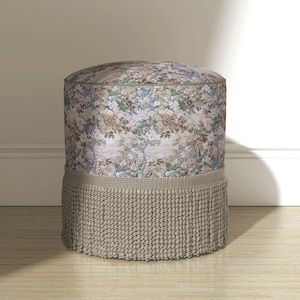 May include: A cylindrical ottoman featuring a multi-colored floral and landscape tapestry fabric upholstery. The base of the ottoman is adorned with a thick, textured beige fringe trim that encircles the entire lower section of the piece.