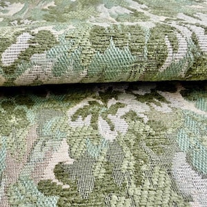 May include: A folded piece of heavy upholstery fabric featuring a dense, woven jacquard pattern in shades of sage green, olive green, and off-white. The textile displays a complex, repeating botanical design with textured, raised weave elements.