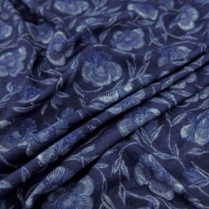May include: A dark blue fabric featuring a repeating floral pattern in a lighter shade of blue. The material is draped, creating soft folds and shadows that highlight the intricate botanical design of flowers and leaves across the surface.