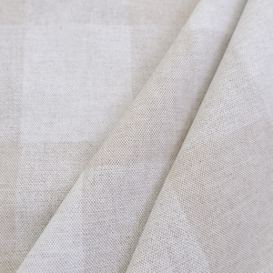 May include: A piece of fabric featuring a subtle, large-scale checkered pattern in shades of light beige and off-white. The material has a visible, textured weave with a matte finish, showing folds that create soft shadows across the surface.