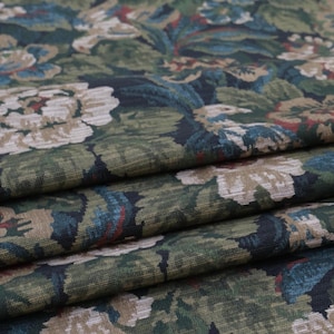 May include: A folded textile fabric featuring a dense floral pattern with large cream-colored blossoms, deep green foliage, and accents of dark blue and muted red. The material has a visible woven texture with multiple horizontal folds across the surface.