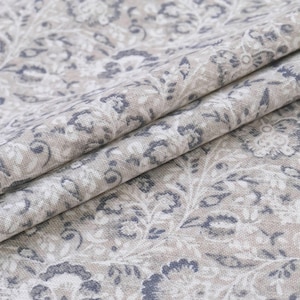 May include: Close-up of folded fabric with a floral pattern in shades of beige, gray, and blue. The fabric has a textured appearance, with visible folds and creases. The design features stylized flowers and leaves.