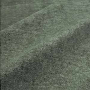 May include: Close-up of a textured fabric in a muted green hue. The fabric has a soft, brushed appearance with subtle variations in color and texture, creating a sense of depth and visual interest. The folds and shadows add to the tactile quality of the material.