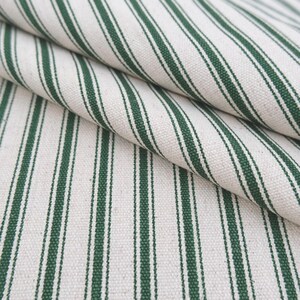 May include: A woven fabric featuring a repeating pattern of dark forest green stripes against an off-white, cream-colored background. The material displays a visible, textured weave with diagonal folds that highlight the vertical striped design.