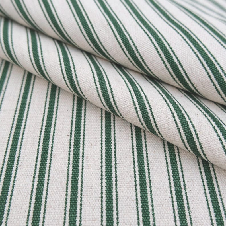 May include: A woven fabric featuring a repeating pattern of dark forest green stripes against an off-white, cream-colored background. The material displays a visible, textured weave with diagonal folds that highlight the vertical striped design.