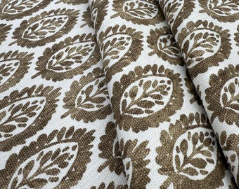 May include: A white fabric featuring a repeating pattern of brown leaf-shaped motifs. Each motif consists of a central leaf design surrounded by a textured, scalloped border. The material has a visible woven texture throughout the entire surface.