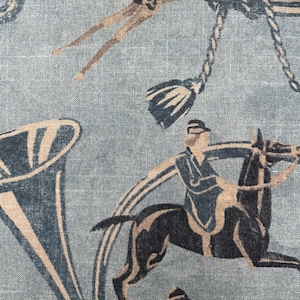 Puede incluir: Primer plano de una tela con un dise&ntilde;o ecuestre repetido. El patr&oacute;n presenta jinetes a caballo, borlas decorativas y formas de cuerno estilizadas en tonos azul marino, beige y marr&oacute;n sobre un fondo azul claro.