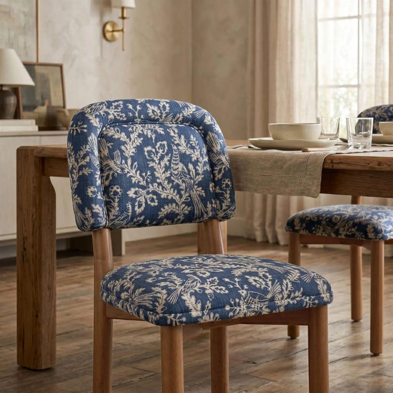 May include: A dining chair with a rounded backrest and a square seat, both upholstered in a blue fabric featuring a white botanical and bird pattern. The chair has a wooden frame with four straight legs, matching the wood of the dining table.