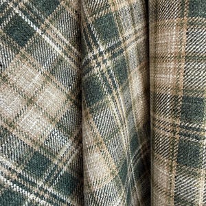 May include: A woven fabric featuring a plaid pattern in shades of forest green, beige, and cream. The textile displays a textured, slightly fuzzy surface with intersecting diagonal lines forming a classic tartan grid across the entire material.
