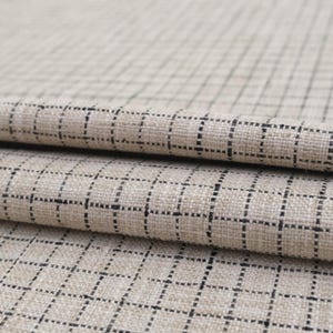 以下が含まれることがあります： 黒い格子模様のベージュ色の生地のクローズアップ。生地は折りたたまれ、テクスチャード加工された素材の層が現れています。格子は均等に配置され、構造化されたデザインを作成しています。生地は天然繊維のようです。