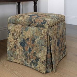 May include: A square upholstered ottoman with a floral pattern in shades of green, brown, and blue. The fabric features a detailed botanical design. The ottoman has a pleated skirt detail and sits on a wooden floor.
