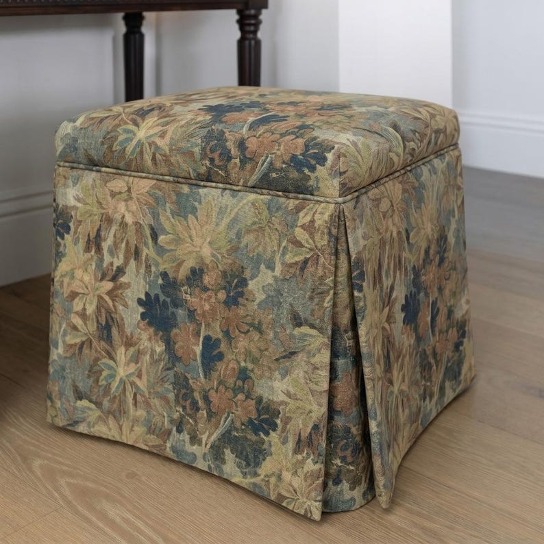 May include: A square upholstered ottoman with a floral pattern in shades of green, brown, and blue. The fabric features a detailed botanical design. The ottoman has a pleated skirt detail and sits on a wooden floor.