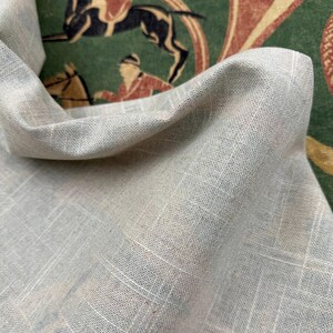 May include: Close-up of a light gray linen fabric with a subtle grid pattern. The fabric is draped, revealing a glimpse of a vintage-style green and red patterned background with equestrian figures. The fabric has a soft, textured appearance.