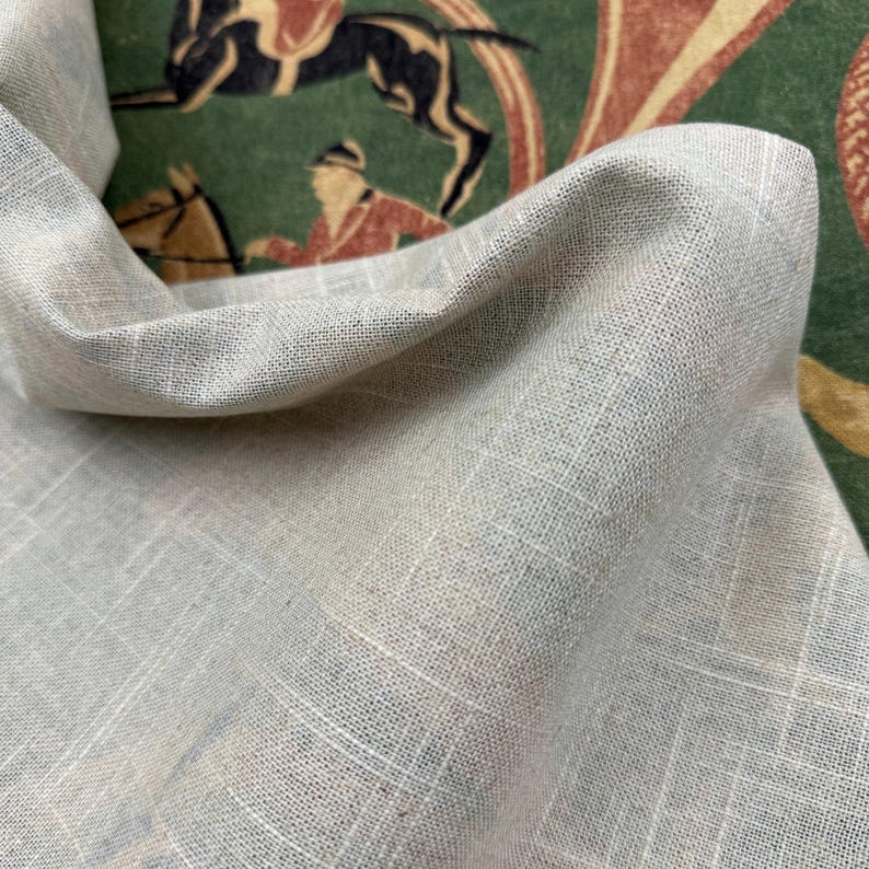 May include: Close-up of a light gray linen fabric with a subtle grid pattern. The fabric is draped, revealing a glimpse of a vintage-style green and red patterned background with equestrian figures. The fabric has a soft, textured appearance.