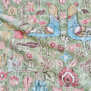 May include: A fabric featuring a repeating pattern of two stylized birds facing each other, rendered in shades of blue, tan, and white. The background is a muted sage green, decorated with intricate floral motifs in pink, yellow, and brown.