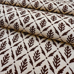 May include: Close-up of a folded fabric with a repeating pattern. The fabric is a light beige color with a dark brown leaf-like design. The pattern is arranged in a grid-like formation, creating a textured appearance. The fabric appears to be a woven material.