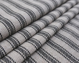May include: A woven fabric featuring a repeating pattern of thick black stripes set against a light beige, textured background. The material is folded in multiple layers, showcasing the visible weave and the consistent vertical alignment of the stripes.
