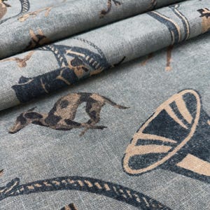 Puede incluir: Primer plano de una tela azul claro con un patr&oacute;n repetido de perros corriendo, cuerdas y otros dise&ntilde;os ecuestres en azul marino y beige. La tela est&aacute; doblada, mostrando textura y profundidad.