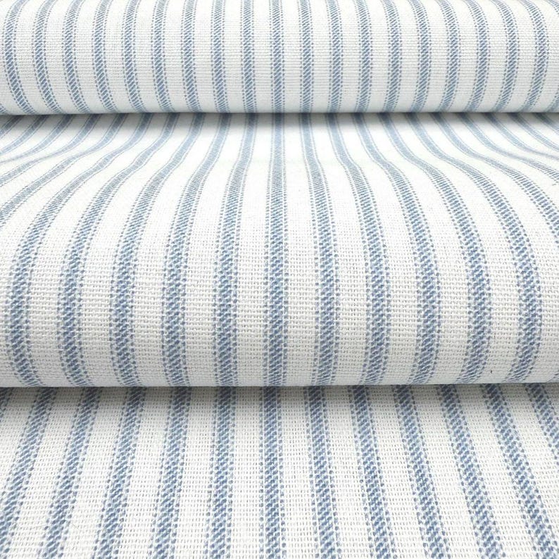 Peut inclure: Un tissu textile pr&eacute;sentant un motif r&eacute;p&eacute;titif de rayures verticales bleues sur un fond blanc. Le mat&eacute;riau poss&egrave;de une texture tiss&eacute;e visible avec une densit&eacute; de tissage constante, montrant plusieurs plis du tissu superpos&eacute;s horizontalement.