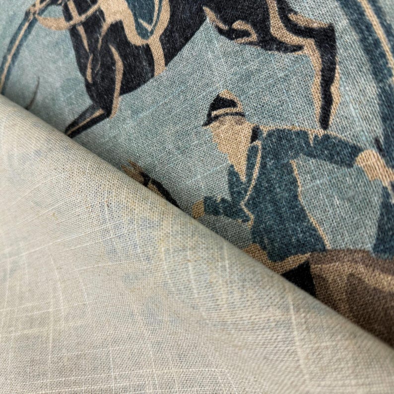 Puede incluir: Primer plano de una tela con un dise&ntilde;o ecuestre vintage. La tela presenta un fondo azul claro con un patr&oacute;n repetido de un caballo y un jinete en tonos azul marino, beige y marr&oacute;n. La tela est&aacute; doblada, revelando un reverso beige claro liso.