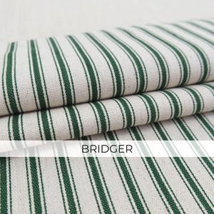 May include: A folded piece of fabric featuring a pattern of dark green vertical stripes on an off-white background. The textile has a visible woven texture and includes the word BRIDGER printed in black capital letters across the center.