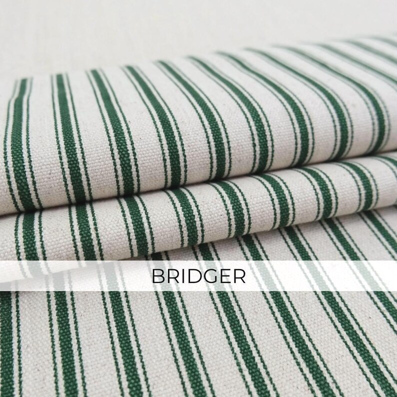 May include: A folded piece of fabric featuring a pattern of dark green vertical stripes on an off-white background. The textile has a visible woven texture and includes the word BRIDGER printed in black capital letters across the center.